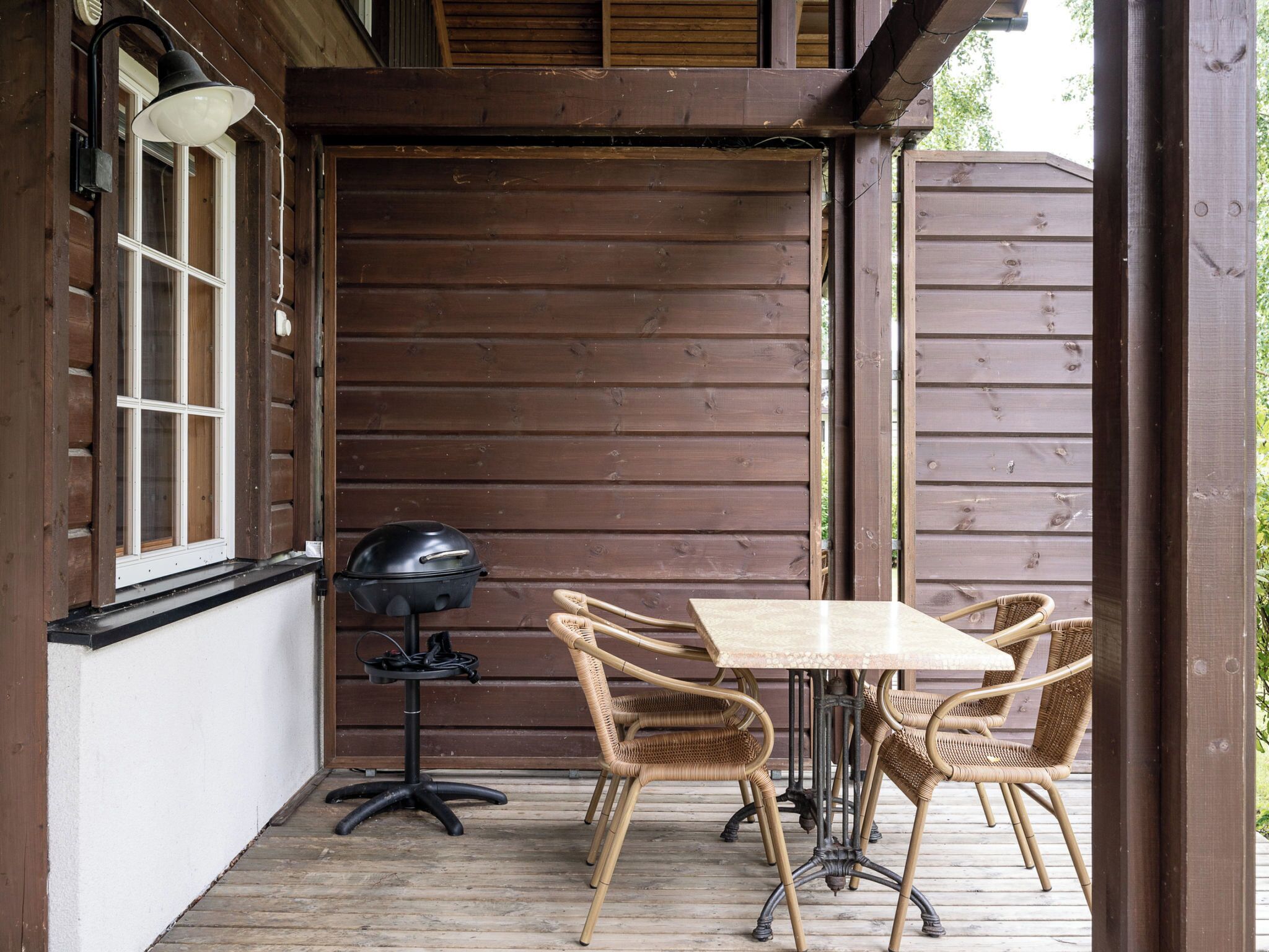 A wooden deck with a table and chairs, a grill, and a window with blinds.