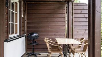 A wooden deck with a table and chairs, a grill, and a window with blinds.