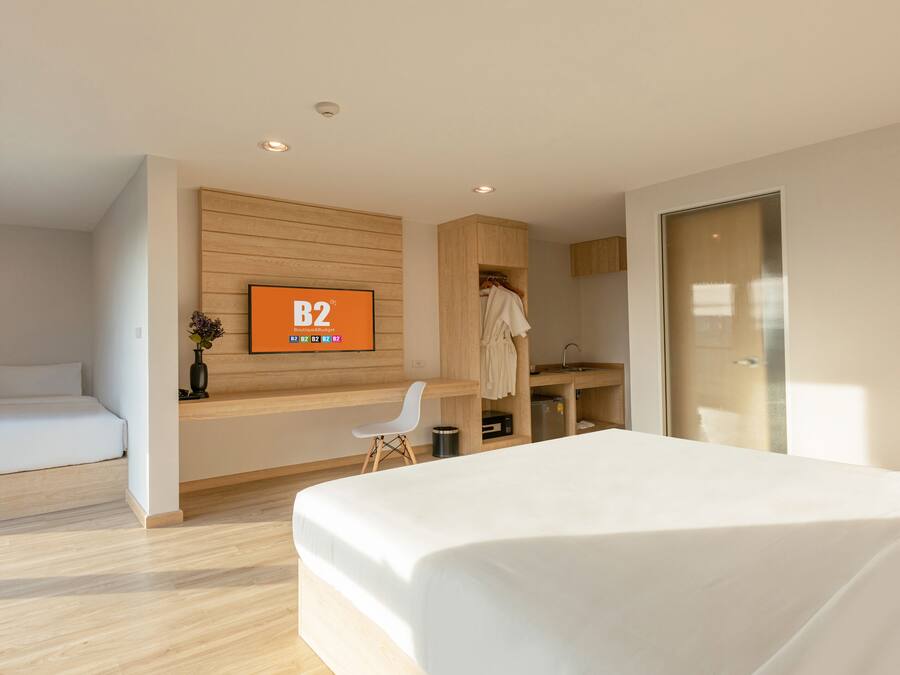 A modern hotel room with a large bed, a smaller bed, a desk, a chair, a robe rack, and a TV displaying 'B2B Boutique'.