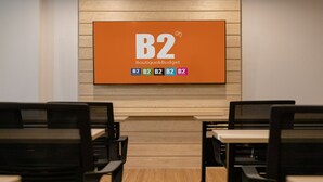 Ein Konferenzraum mit einer großen Leinwand, auf der die Logos 'B2 Boutique&Budget' und 'B2 Premier' zu sehen sind. Zwei schwarze Stühle sind auf den Bildschirm gerichtet.