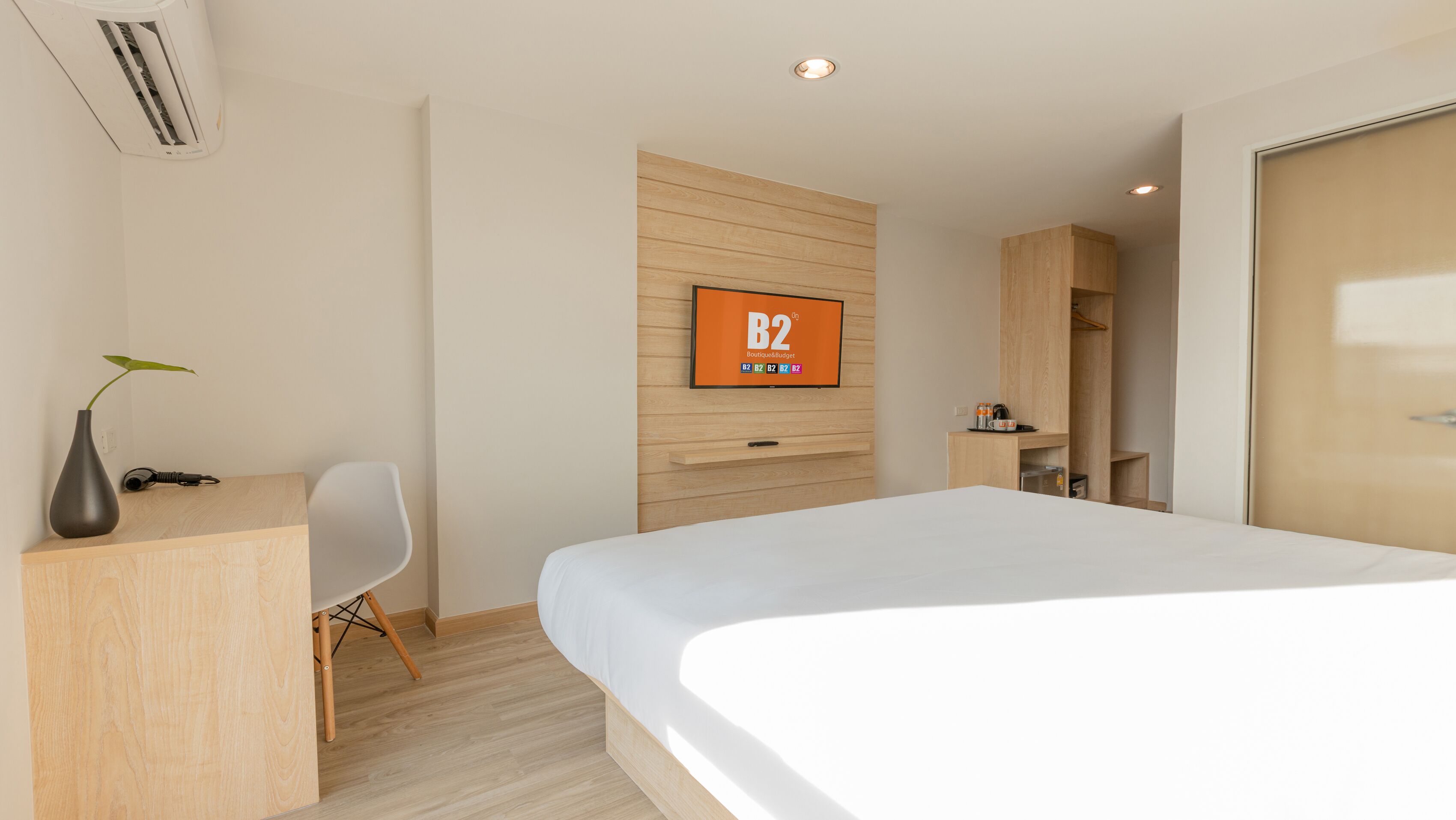 A modern hotel room with a bed, a desk, a chair, a TV mounted on the wall, and a wardrobe.