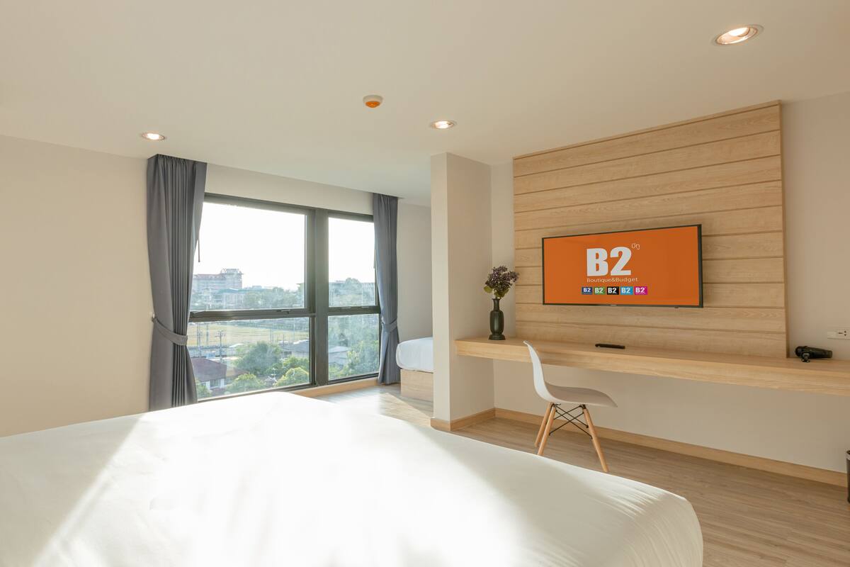 A hotel room with a large bed, a desk with a computer, a TV on the wall, and a window with a city view.