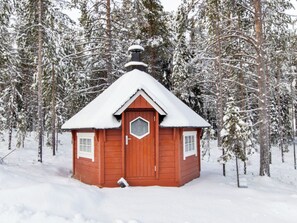 A small, single-story wooden cabin with a snow-covered roof, surrounded by a forest.