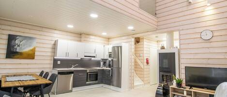 Une cuisine moderne équipée d’appareils électroménagers en acier inoxydable, un coin repas avec une table et des chaises en bois, et un salon avec un téléviseur et des fauteuils.