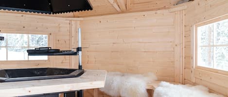 Interior de una cabaña de madera con una mesa, una parrilla negra y una ventana que da a árboles cubiertos de nieve.