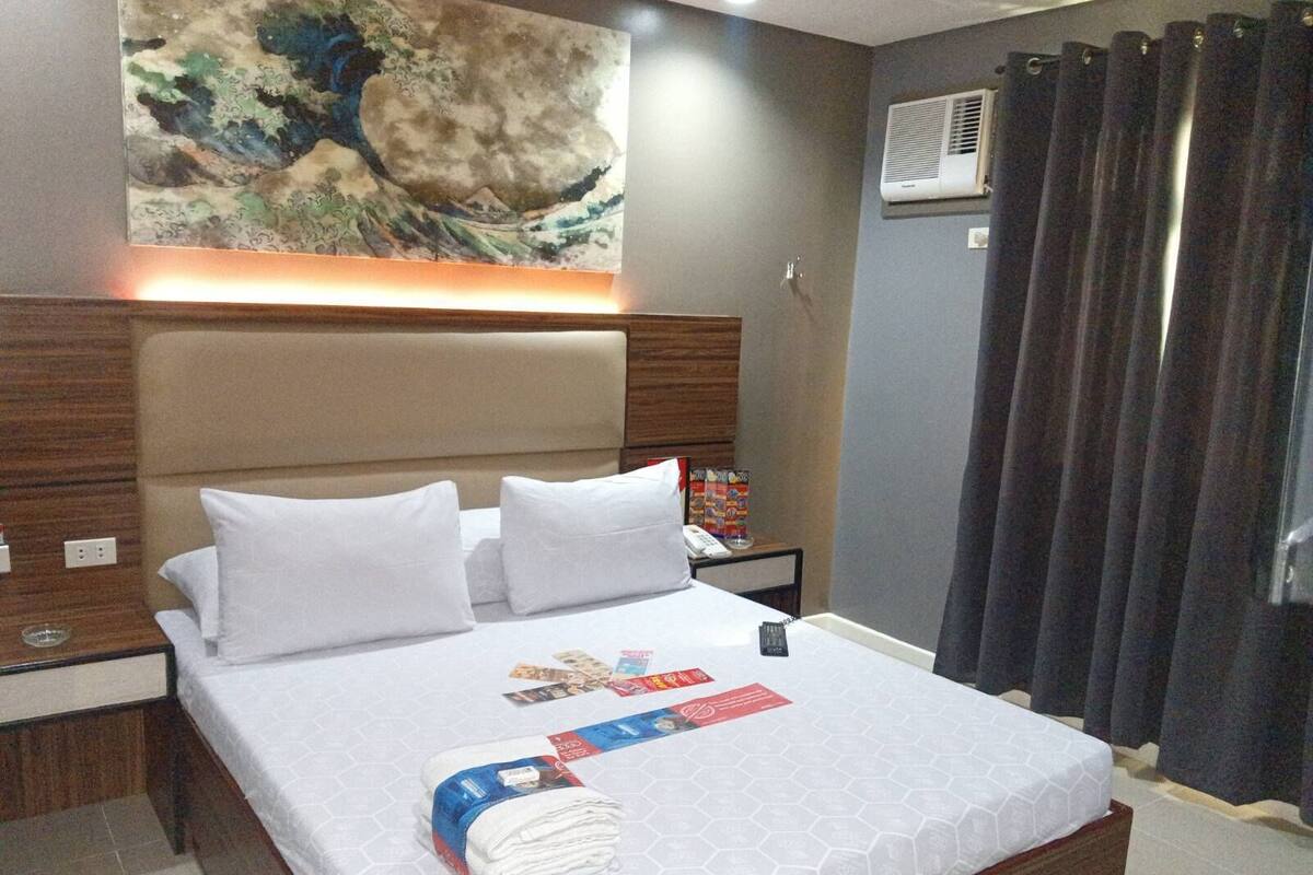 Ein Hotelzimmer mit einem großen Bett, einem Kopfteil, einem Nachttisch mit Telefon und Zeitschrift sowie einer Wand-Klimaanlage und einem Vorhang.
