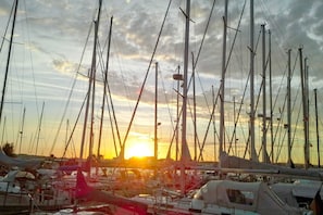 Un port de plaisance où sont amarrés de nombreux voiliers, un ciel dégagé et le soleil qui se couche à l’horizon.