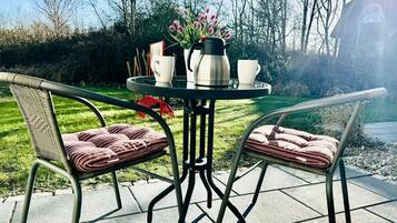 Mesa redonda con dos sillas, una cafetera y dos tazas en un patio con césped y árboles.