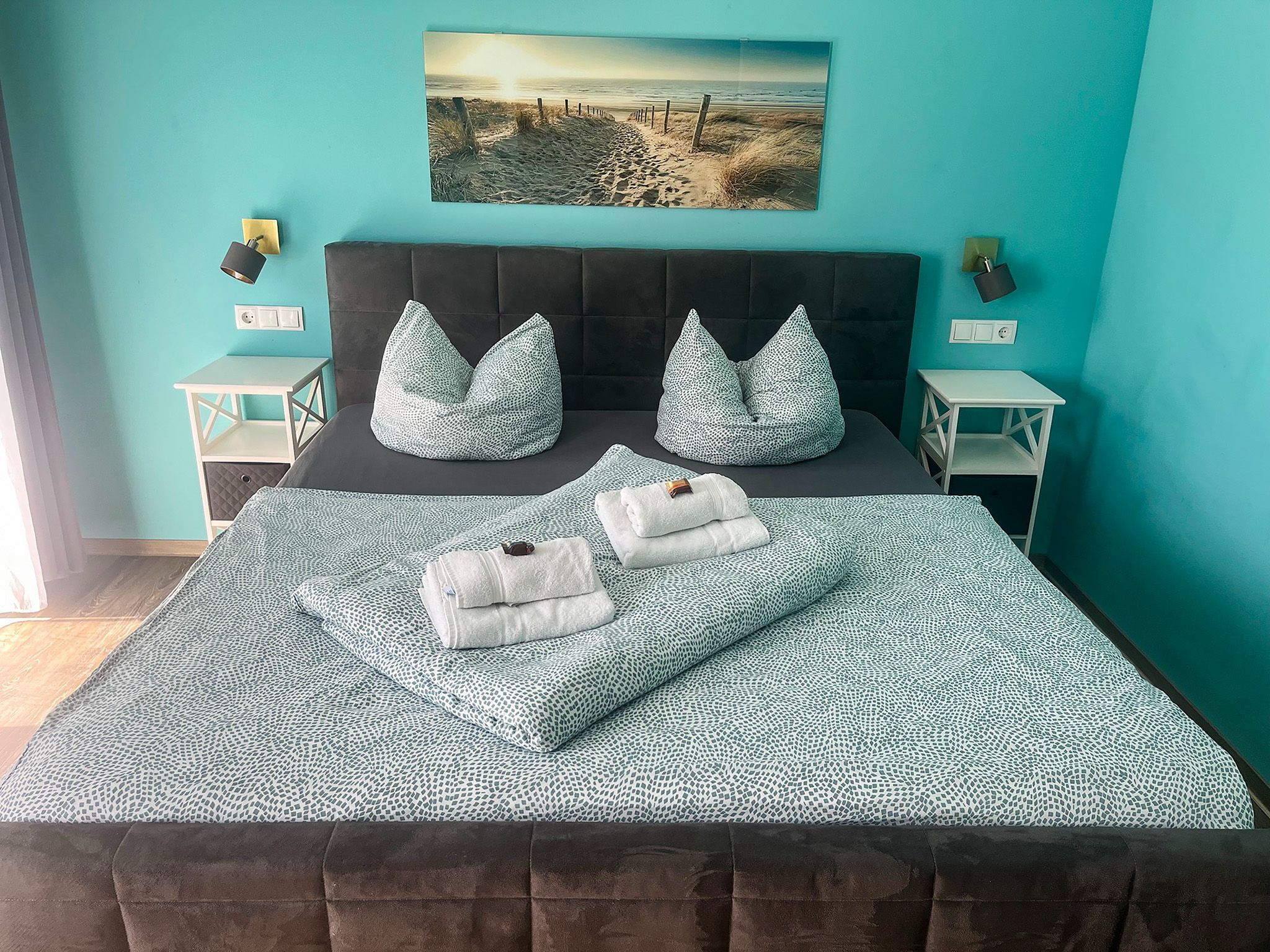 A bedroom with a bed, two nightstands, a wall-mounted lamp, a painting, and a view of the beach.