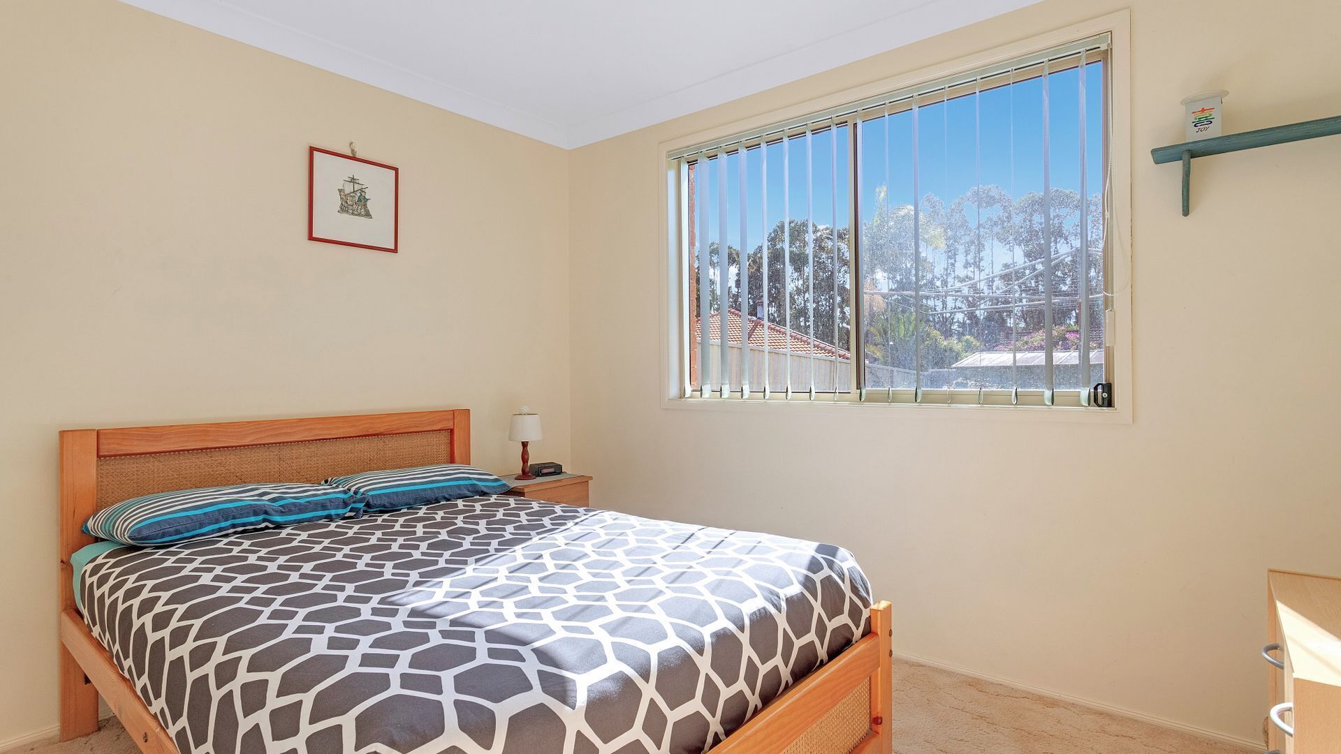 A bedroom with a bed, a nightstand, a lamp, a window with blinds, and a framed picture on the wall.