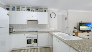A modern kitchen with white cabinetry, granite countertops, a built-in oven, a sink, and a television on a wooden stand.