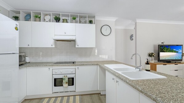 Une cuisine moderne avec des meubles blancs, des plans de travail en granit, un four encastré, un évier et un téléviseur posé sur un meuble en bois.