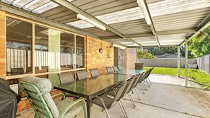Une terrasse couverte avec une longue table et des chaises, un mur en briques et une véranda en verre donnant sur le jardin.