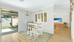A room with a wooden floor, a white table with stools, a large window with blinds, and a TV on a wooden cabinet.