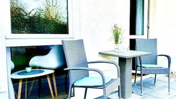 Un área de estar al aire libre con sillas de mimbre y una mesa, una ventana con vista a árboles, y una puerta.