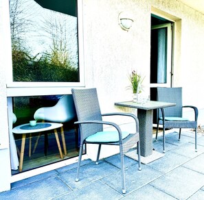 Un espace extérieur aménagé avec des chaises et une table en osier, une fenêtre donnant sur les arbres et une porte.
