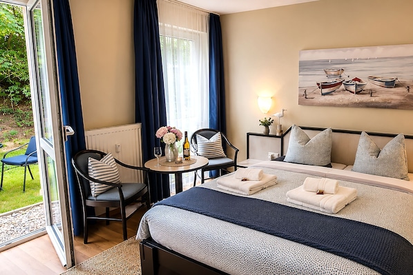 A hotel room with a bed, a table with a vase and wine bottle, a chair, and a painting on the wall.