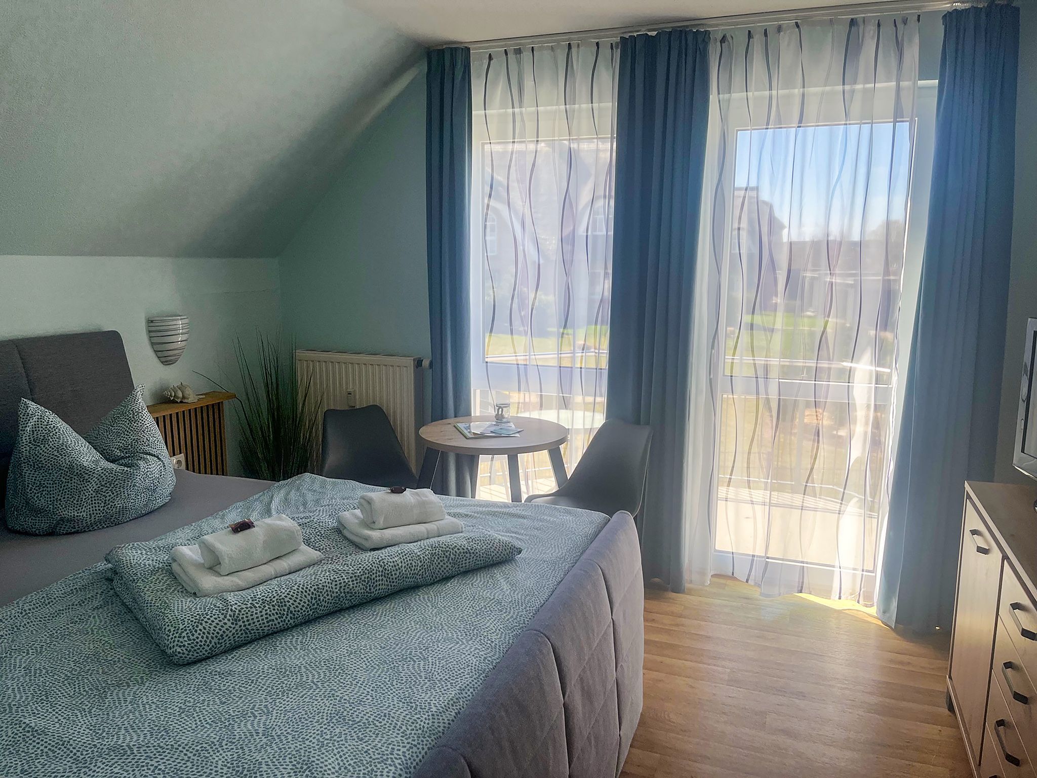 Quarto com cama, uma mesinha, cadeira, radiador e vista pela janela.