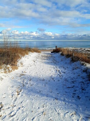A snow-covered path leading to a beach with a clear view of the ocean and a cloudy sky.