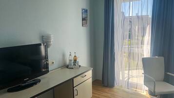 Ein Hotelzimmer mit Flachbildfernseher, einem kleinen Tisch mit Flaschen, einem Sessel und einem Fenster mit Vorhängen.