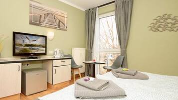 Quarto de hotel com cama, escrivaninha com televisão, pequeno frigorífico, cadeira e janela com cortinas.