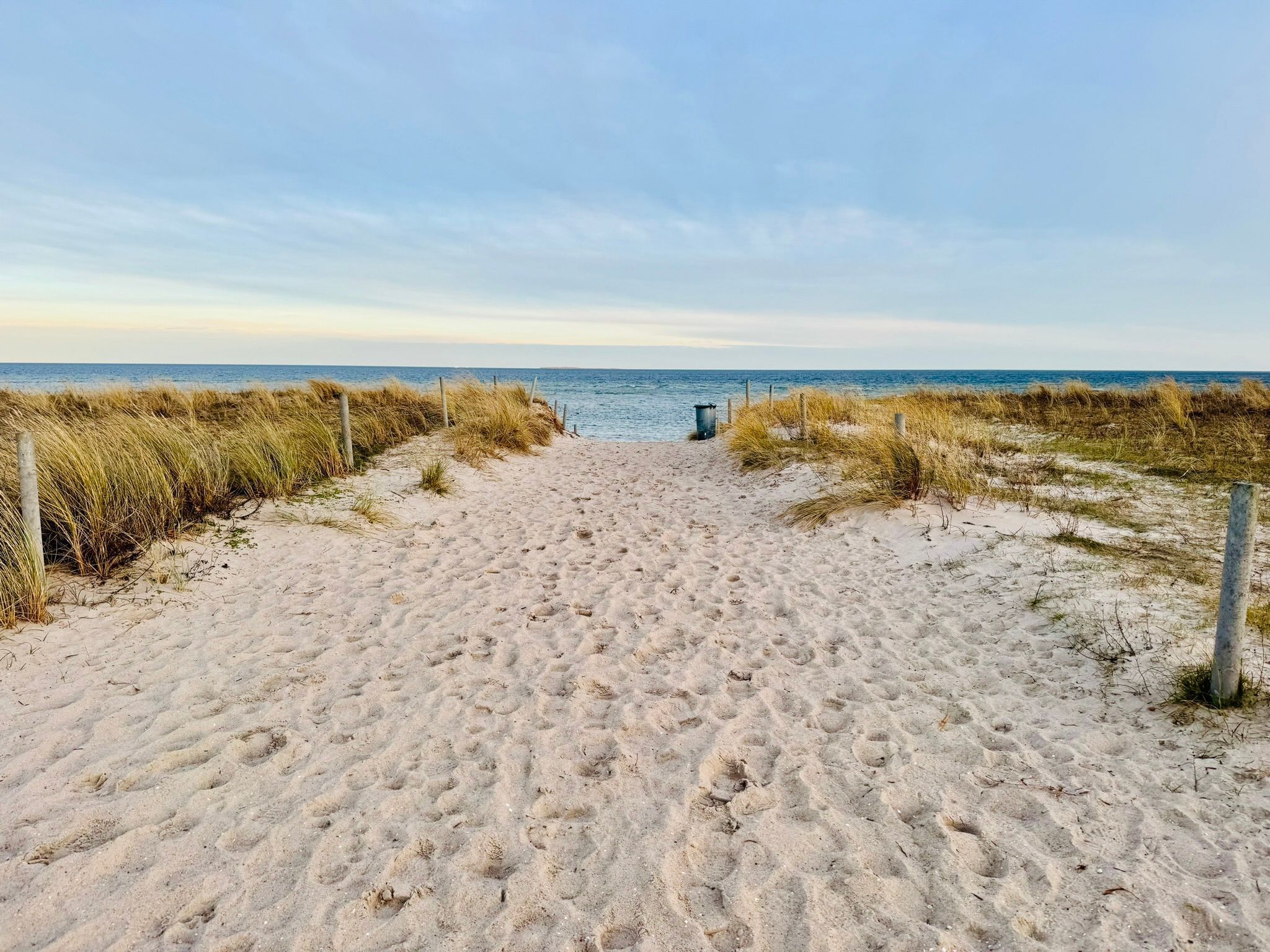 Ein sandiger Küstenpfad führt zum Meer, gesäumt von Gras und Holzbalken.