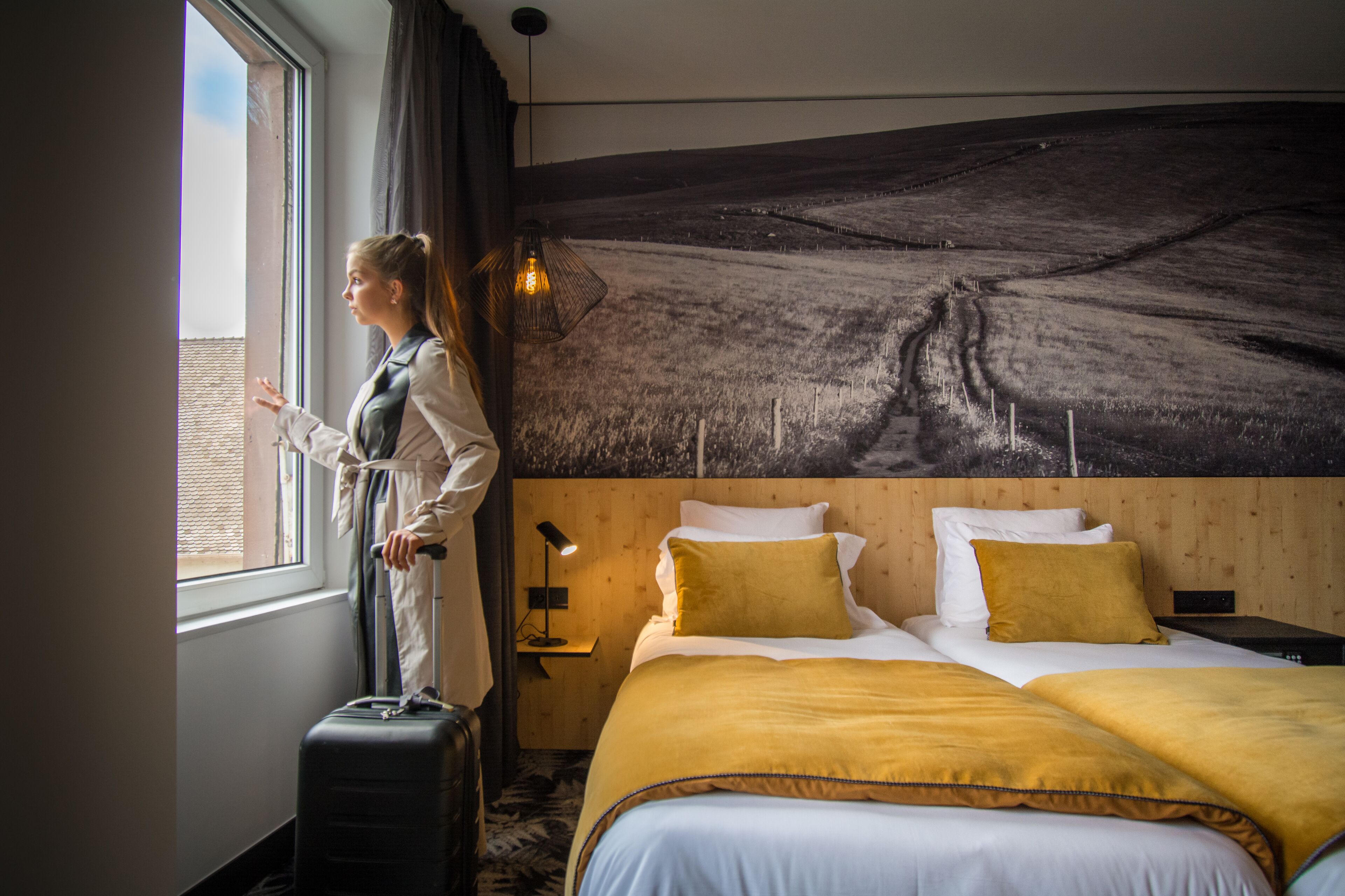 Una habitación de hotel con una cama grande, una ventana con cortinas, una lámpara de pared y una maleta.