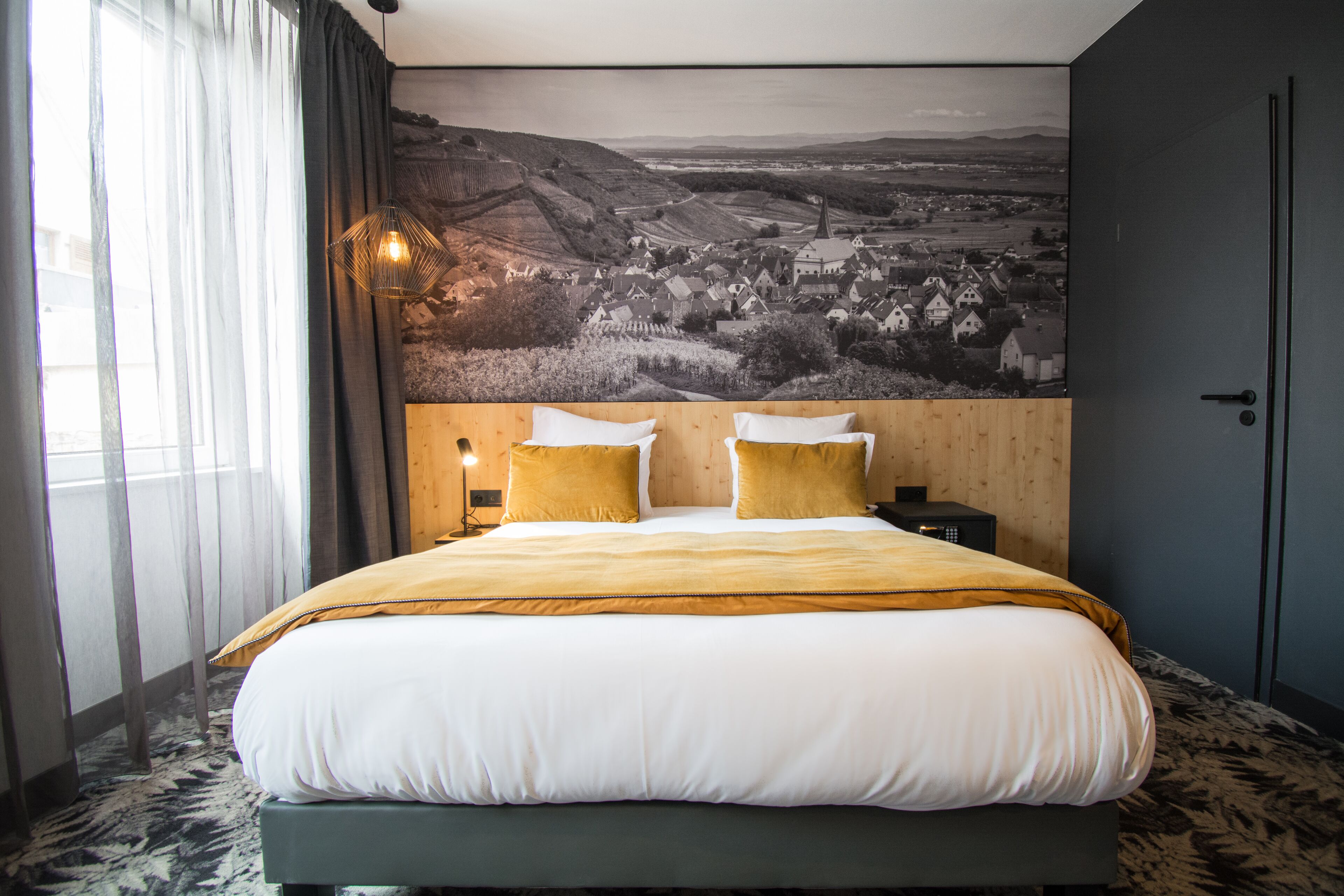Camera d'albergo con un letto grande, una testiera in legno, un murale con una veduta urbana, una finestra con tende leggere e un comodino con una lampada.
