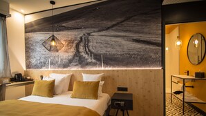 A hotel room with a large bed, a nightstand, a small table, a mirror, and a wall-mounted artwork depicting a rural landscape. - L'Hôtel & Spa  Ribeauvillé (Ribeauvillé)