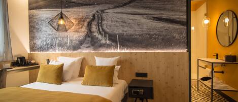 A hotel room with a large bed, a nightstand, a small table, a mirror, and a wall-mounted artwork depicting a rural landscape.