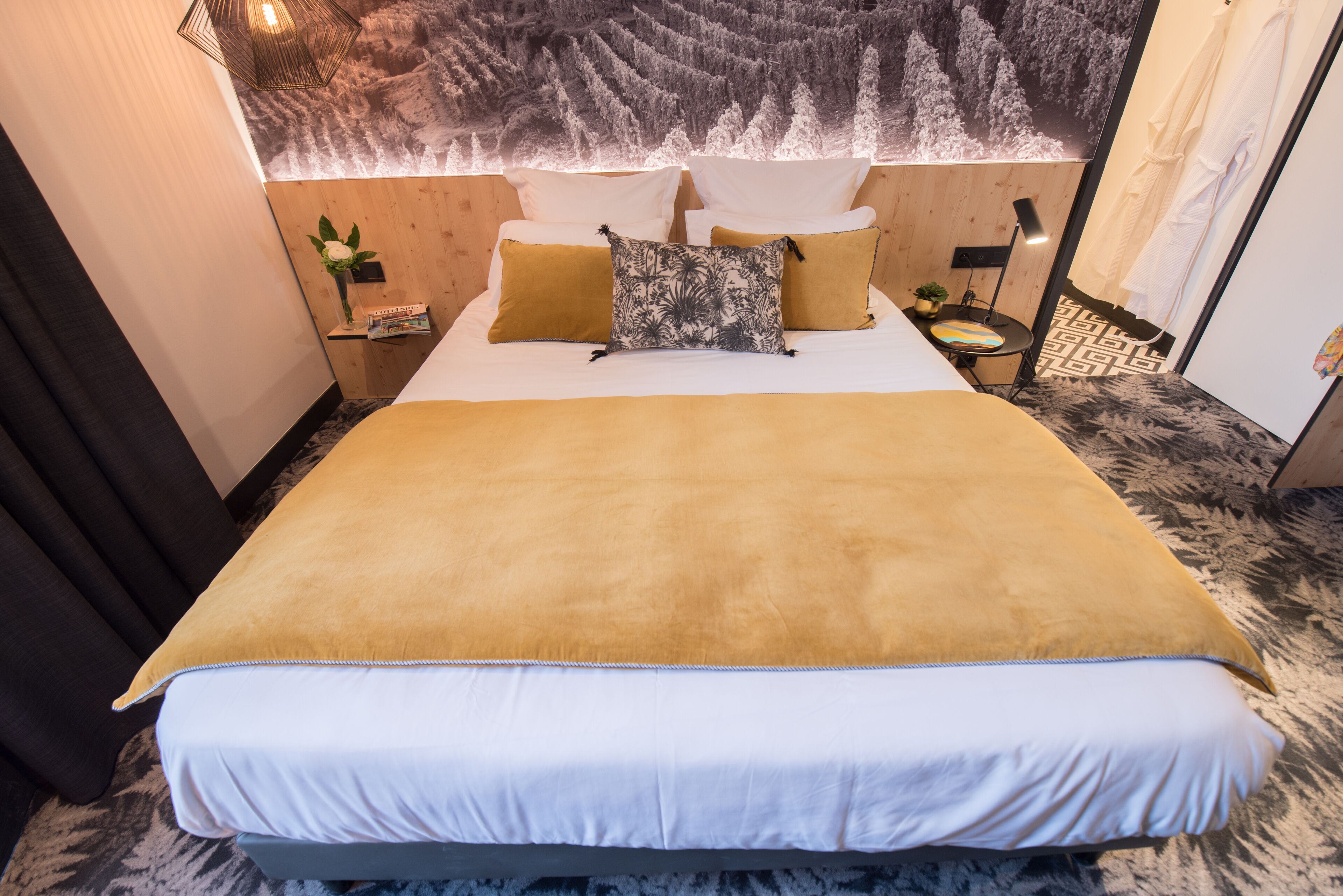 A neatly made bed with a white and yellow bedspread, a headboard with a forest-themed wall art, and a small bedside table with a lamp.