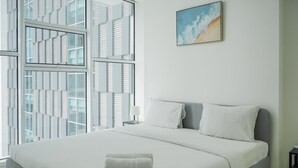 A modern hotel room with a large bed, white bedding, a grey headboard, and a view of a cityscape through floor-to-ceiling windows. - Fully Furnished with Modern Design 1BR Brooklyn Apartment (South Tangerang)