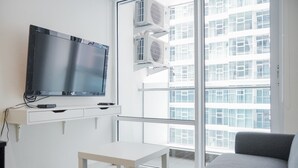 A modern living room with a flat-screen TV mounted on a white wall, a grey sofa, a small white table, and a large window with a view of a high-rise building. - Fully Furnished with Modern Design 1BR Brooklyn Apartment (South Tangerang)