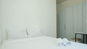 A neatly made bed with white linens and pillows, a small table with a chair, and a wardrobe with a light wood finish. - Cozy with River View @ 2BR Teluk Intan Apartment (Jakarta)
