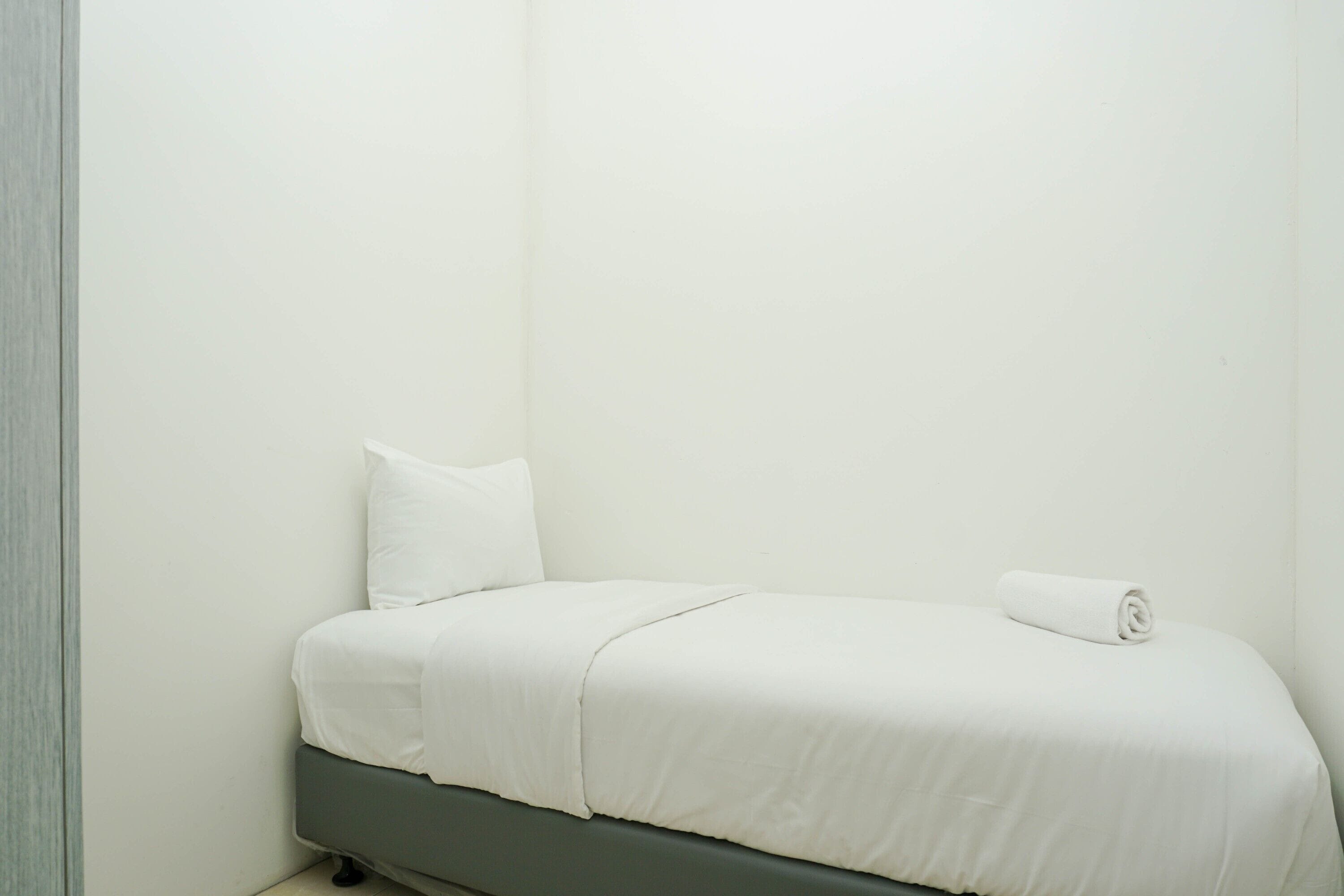 Quarto pequeno com uma cama de solteiro, cabeceira verde, roupa de cama branca e uma toalha enrolada na cama.