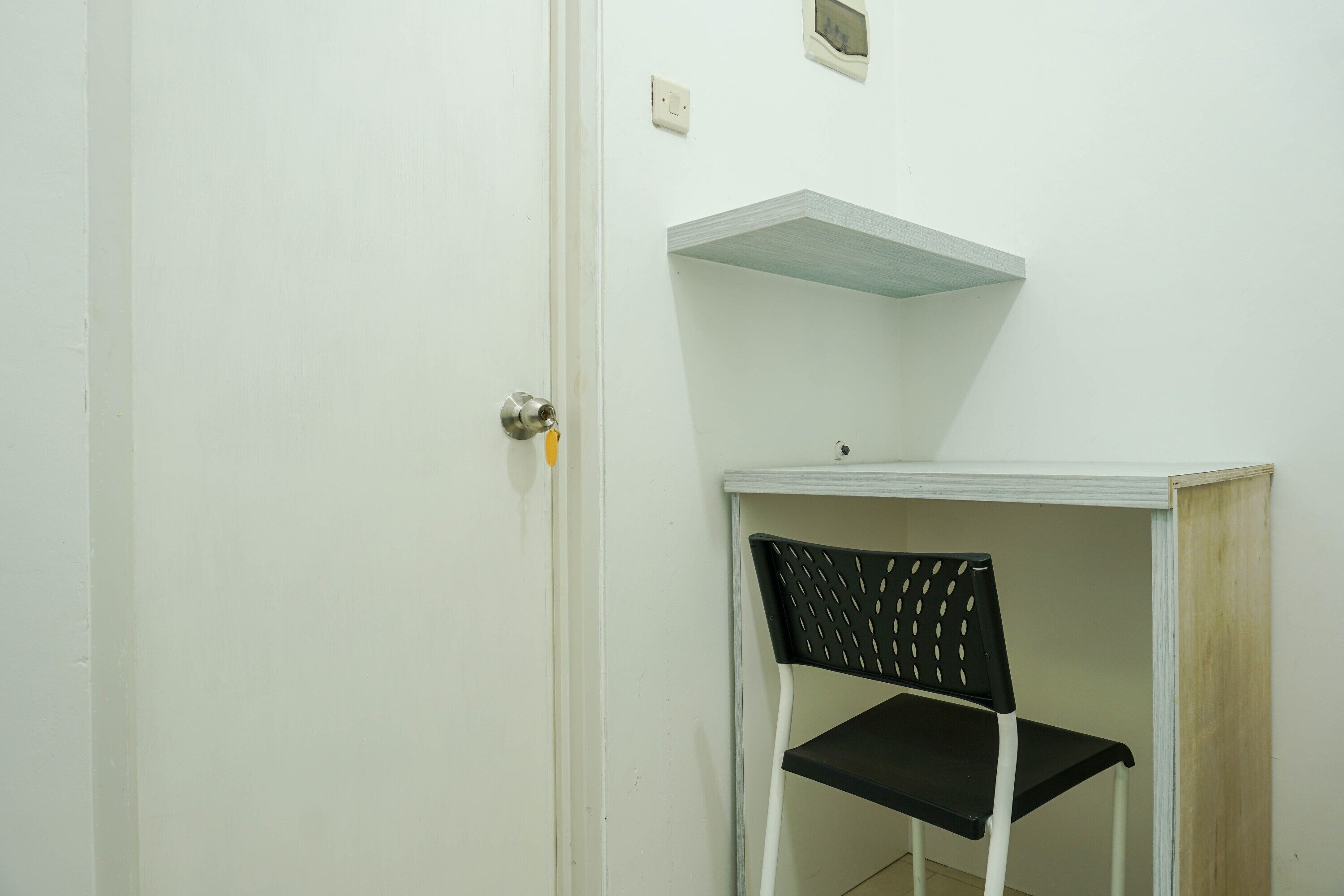A white door with a silver handle, a black chair with a perforated backrest, and a white shelf above the chair.