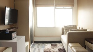 Une chambre d’hôtel moderne avec un téléviseur à écran plat posé sur une console blanche, un canapé beige, une petite table de chevet et une grande fenêtre avec des stores.