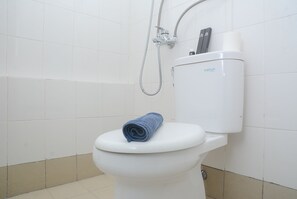 Uma casa de banho branca com uma toalha azul no assento e um chuveiro fixado à parede.