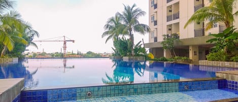 A swimming pool with blue tiles, surrounded by palm trees and a multi-story building.