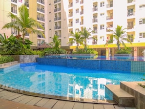 Une piscine aux carreaux bleus, entourée de verdure et bordée d’un immeuble à plusieurs étages doté de balcons.