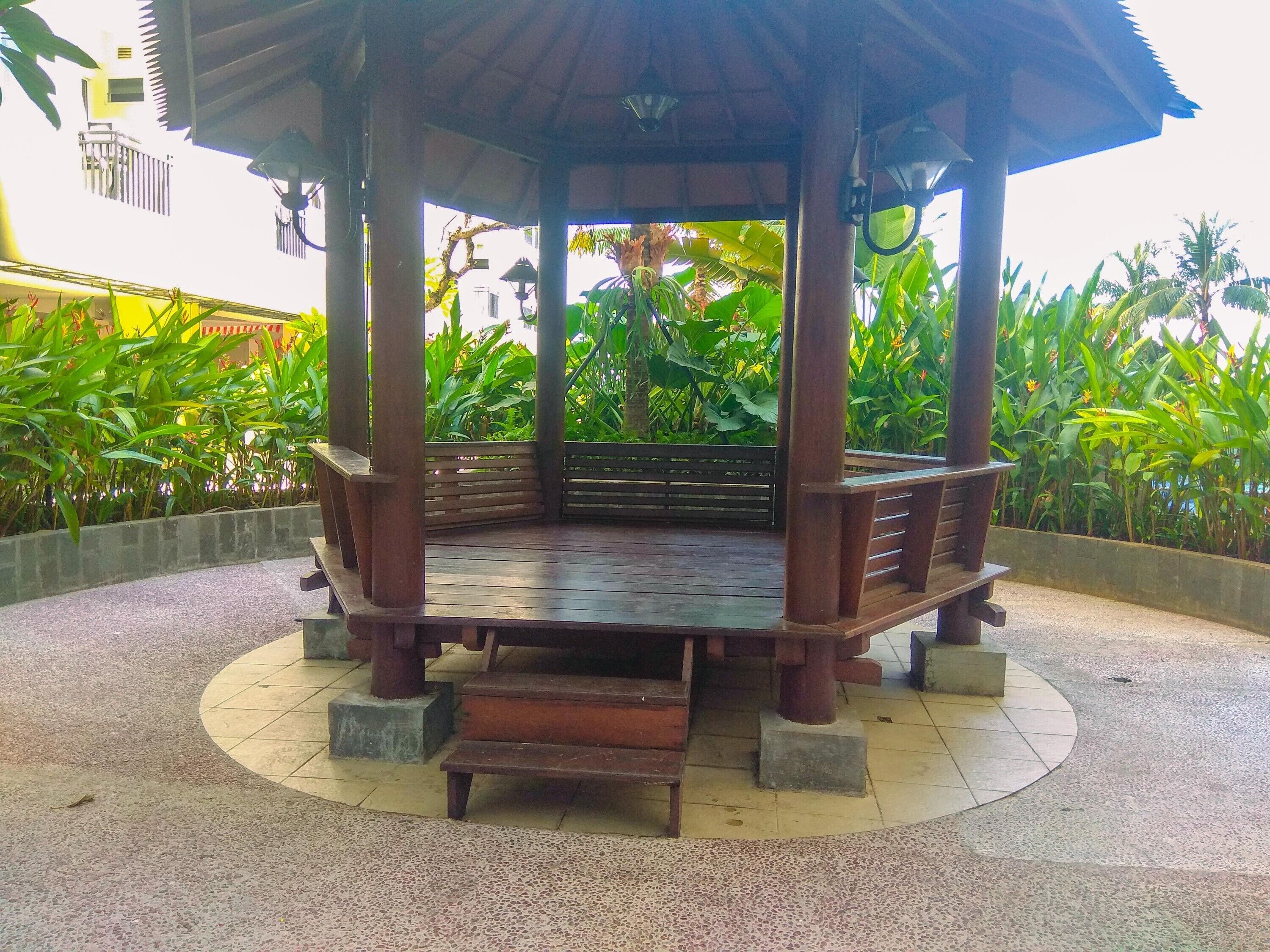 A wooden gazebo with a bench, surrounded by a circular tiled floor, and lush green plants in the background.