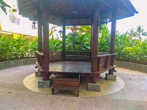 Un belvédère en bois avec un banc, entouré d’un sol circulaire recouvert de carreaux, et une végétation luxuriante en arrière-plan.