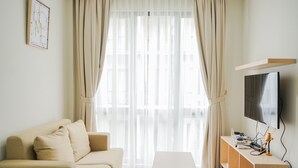 A living room with a beige sofa, a small white table, a TV on a wooden stand, and sheer curtains. - Elegant and Relaxing 1BR Asatti Apartment BSD (Pagedangan)