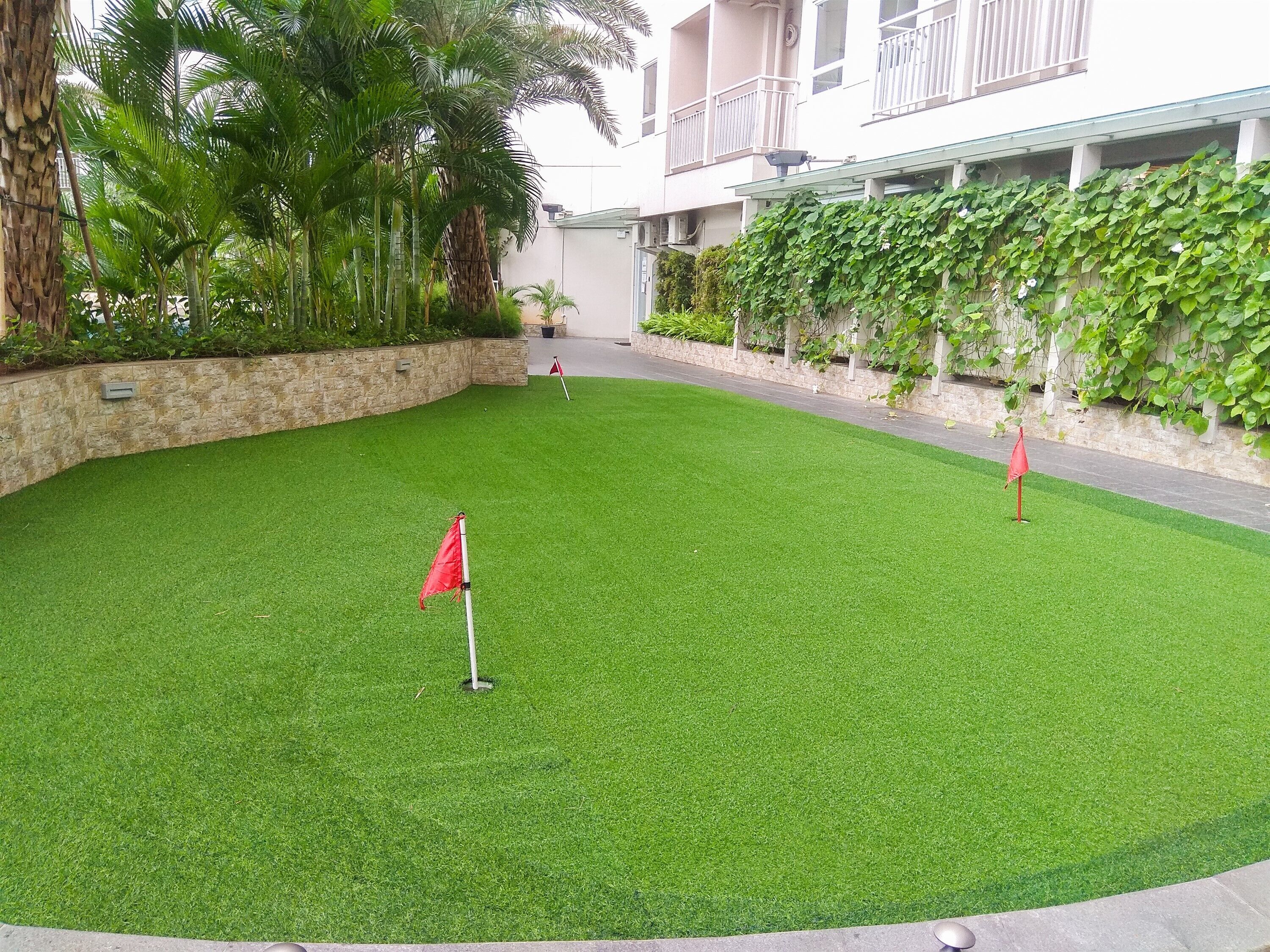 A well-maintained artificial grass area with a red flag marker, surrounded by stone walls and lush greenery.