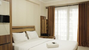 A hotel room with a wooden headboard, a flat-screen TV mounted on the wall, a bedside table with a lamp, a large window with curtains, and a neatly made bed with a rolled towel. - Spacious Studio Room at Gateway Pasteur Apartment near Exit Toll Pasteur (Cimahi)