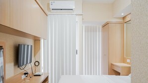 A modern hotel room with a bed, a desk with a flat-screen TV, a wall-mounted air conditioner, and a window with blinds. - Pool View Studio Apartment at M-Town Residence (Gading Serpong)