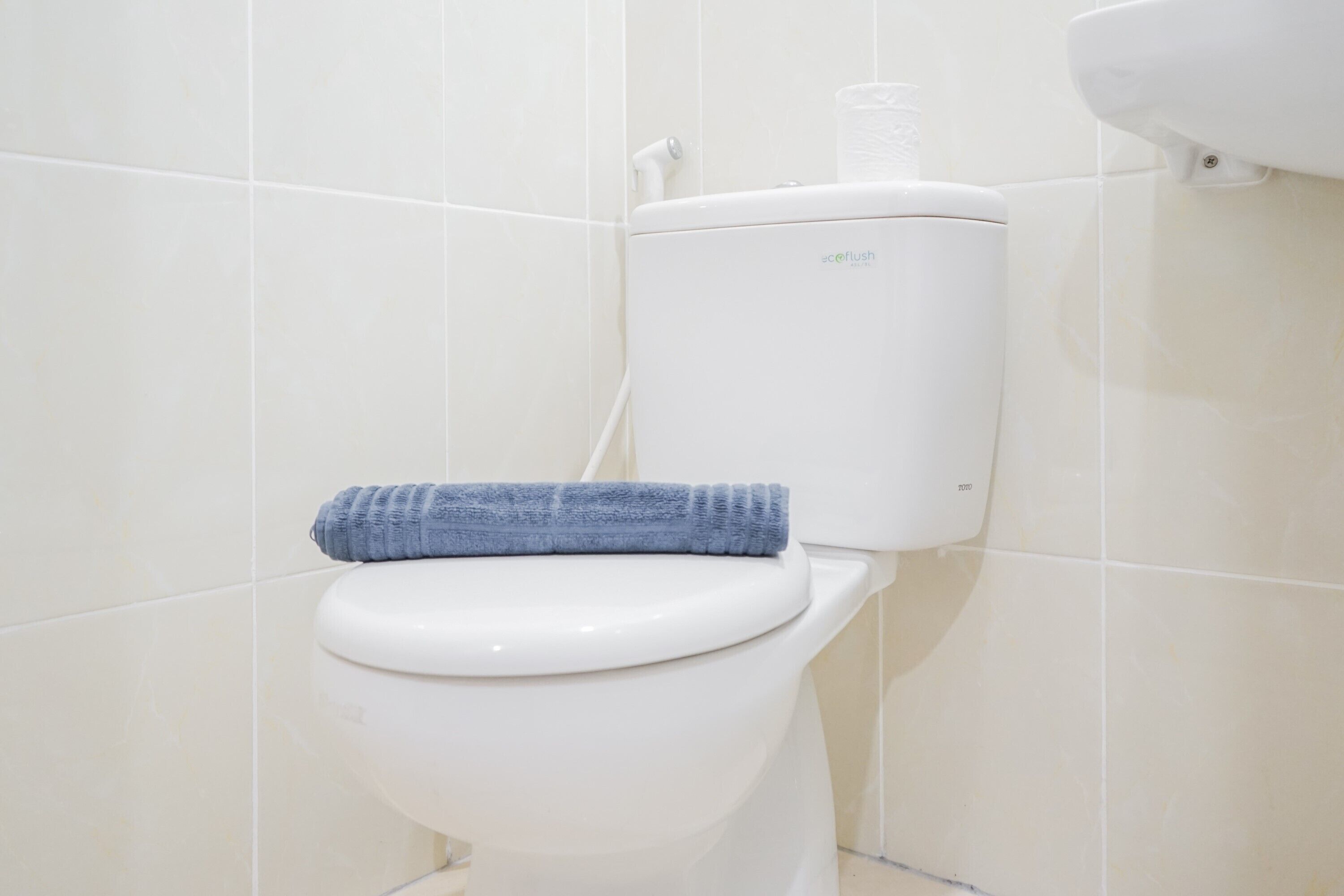 A toilet with a blue towel on the seat and a water tank labeled "ecoflush" in a bathroom with white tiled walls.