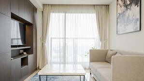 Ein modernes Wohnzimmer mit Fernseher, einem beigen Sofa, einem weißen Couchtisch und einem großen Fenster mit durchsichtigen Vorhängen.