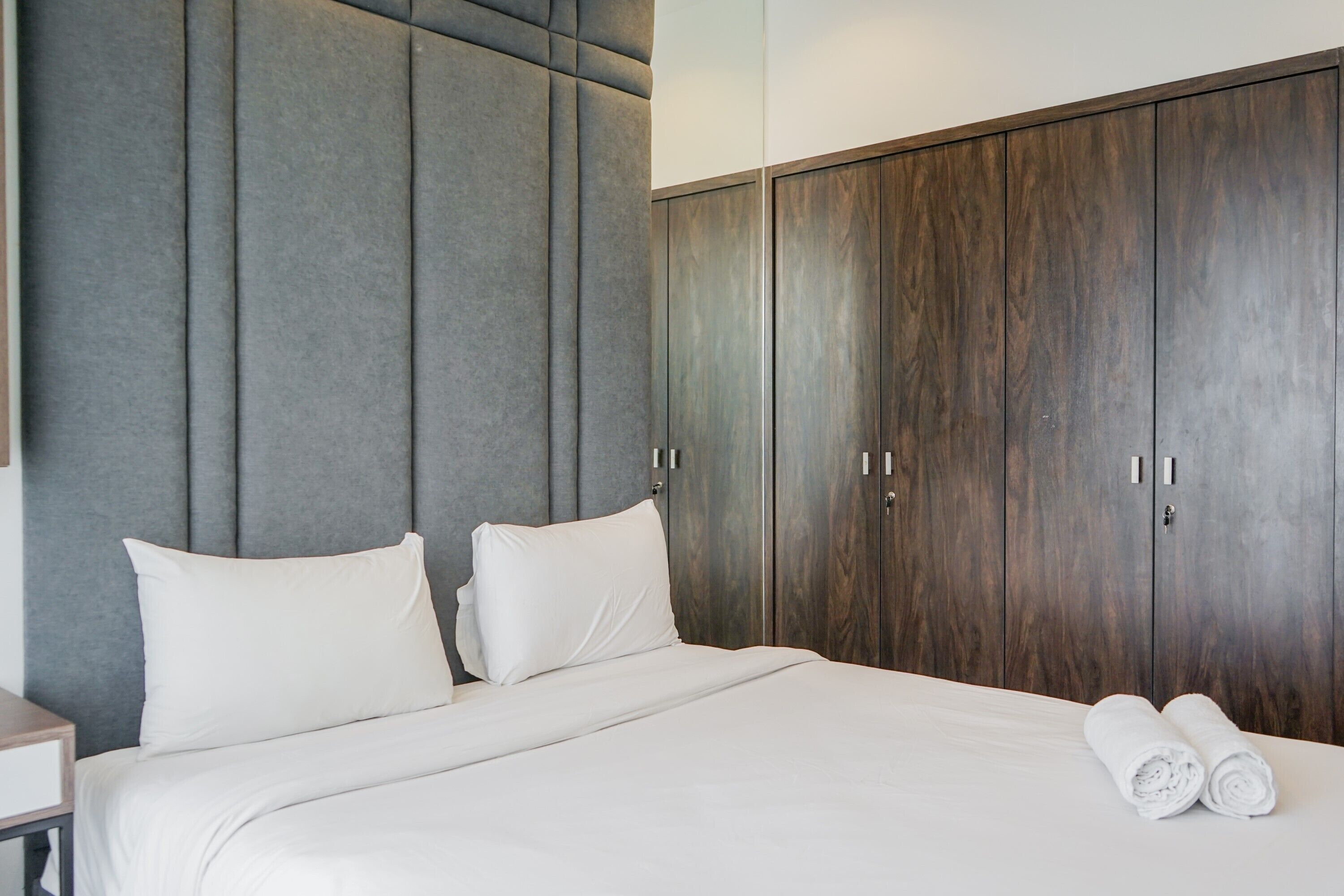 A hotel room with a large bed, grey padded headboard, wooden wardrobe, and white linens.