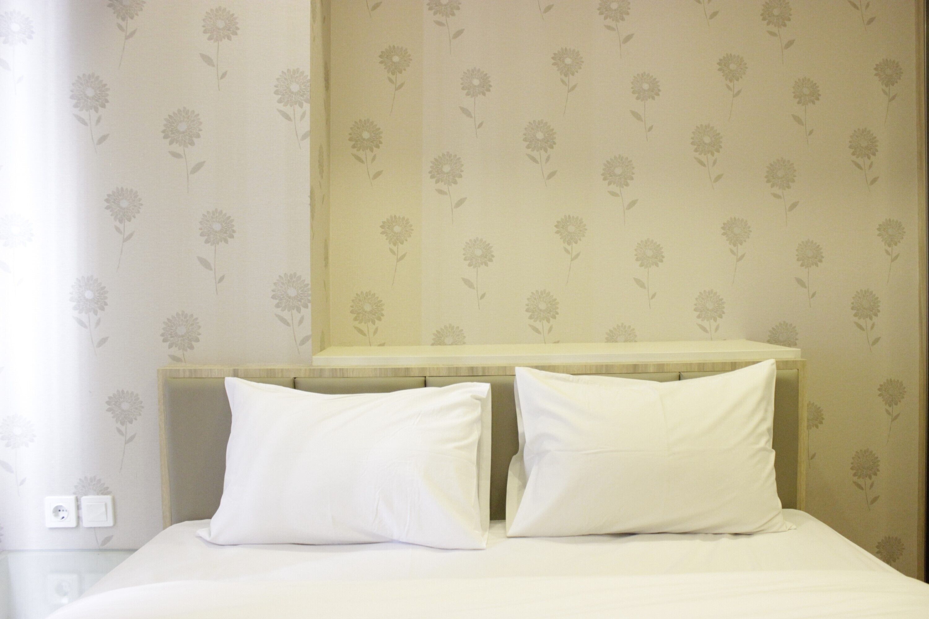 A hotel room with a bed, two white pillows, a wooden headboard, and floral wallpaper.